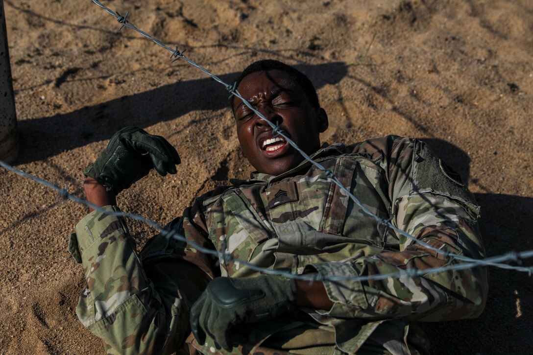 A soldier maneuvers in the dirt under a wire obstacle.