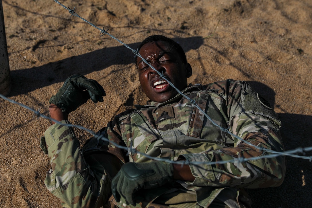 A soldier maneuvers in the dirt under a wire obstacle.