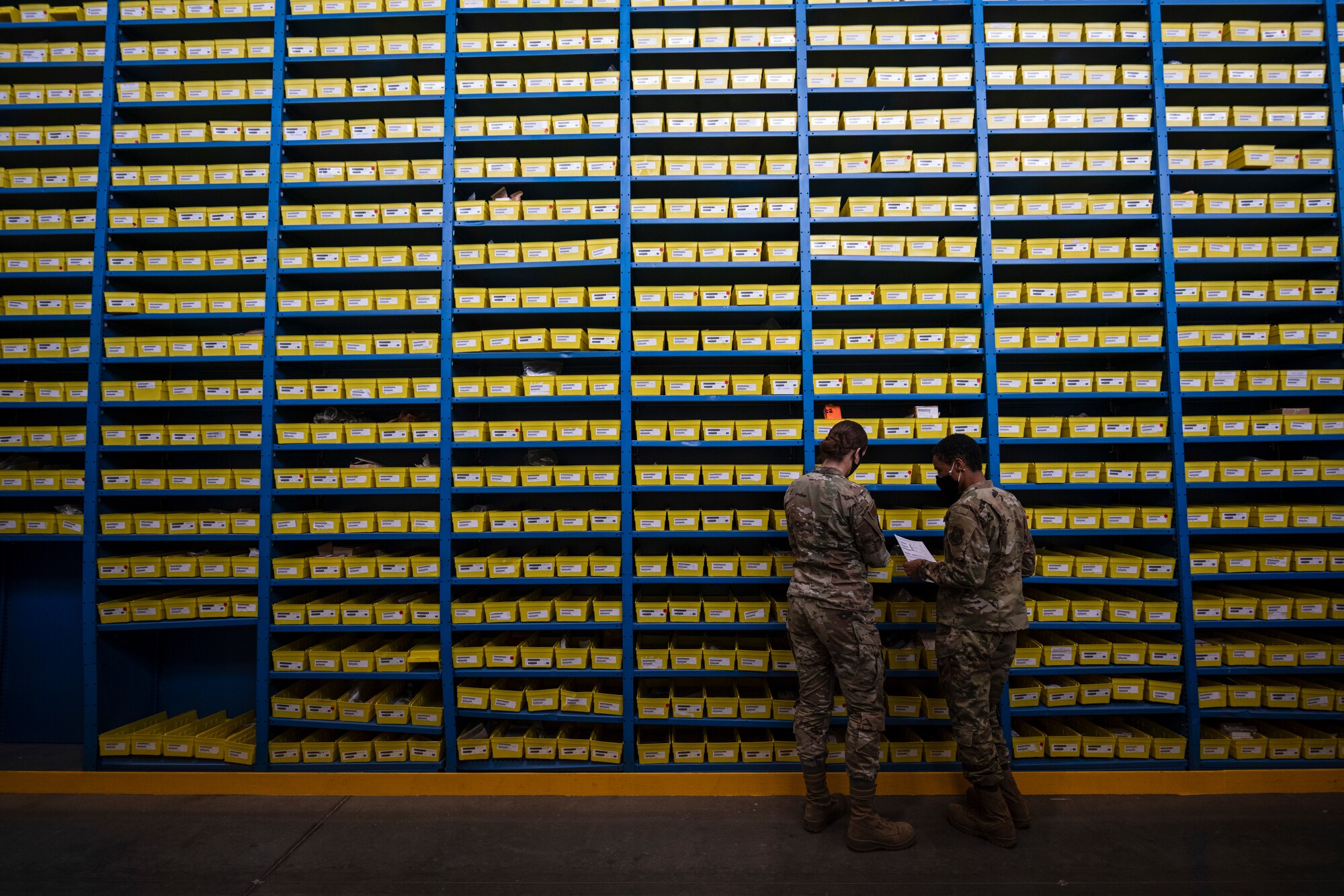 Two female Airmen stand next to each other next to a very tall wall of shelves