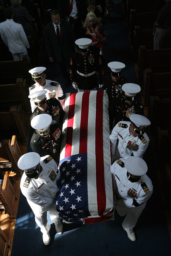 Eight pallbearers dressed in military uniforms carry a coffin draped with an American flag.