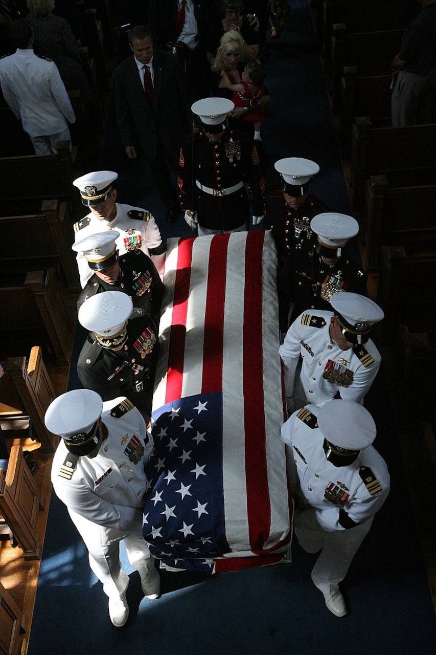 Eight pallbearers dressed in military uniforms carry a coffin draped with an American flag.
