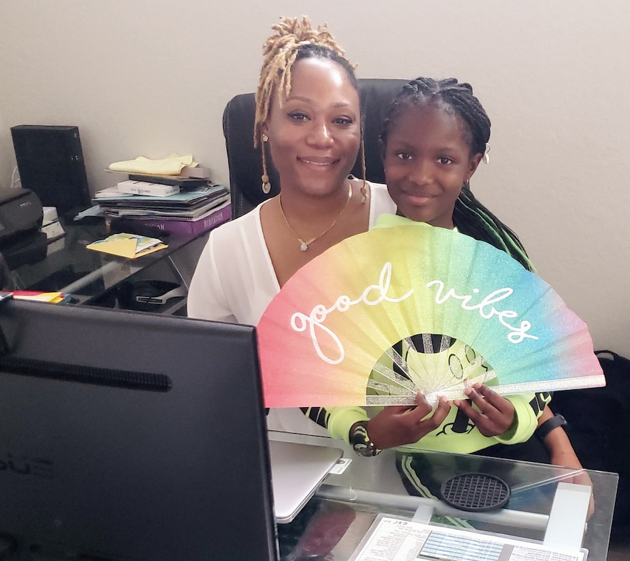 daughter sits on mothers lap in home office displaying a rainbow fan with good vibes written on it