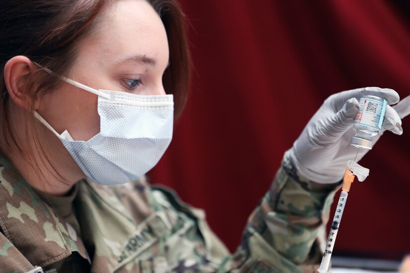 A soldier wearing a face mask and gloves holds a vial while inserting a syringe into it.