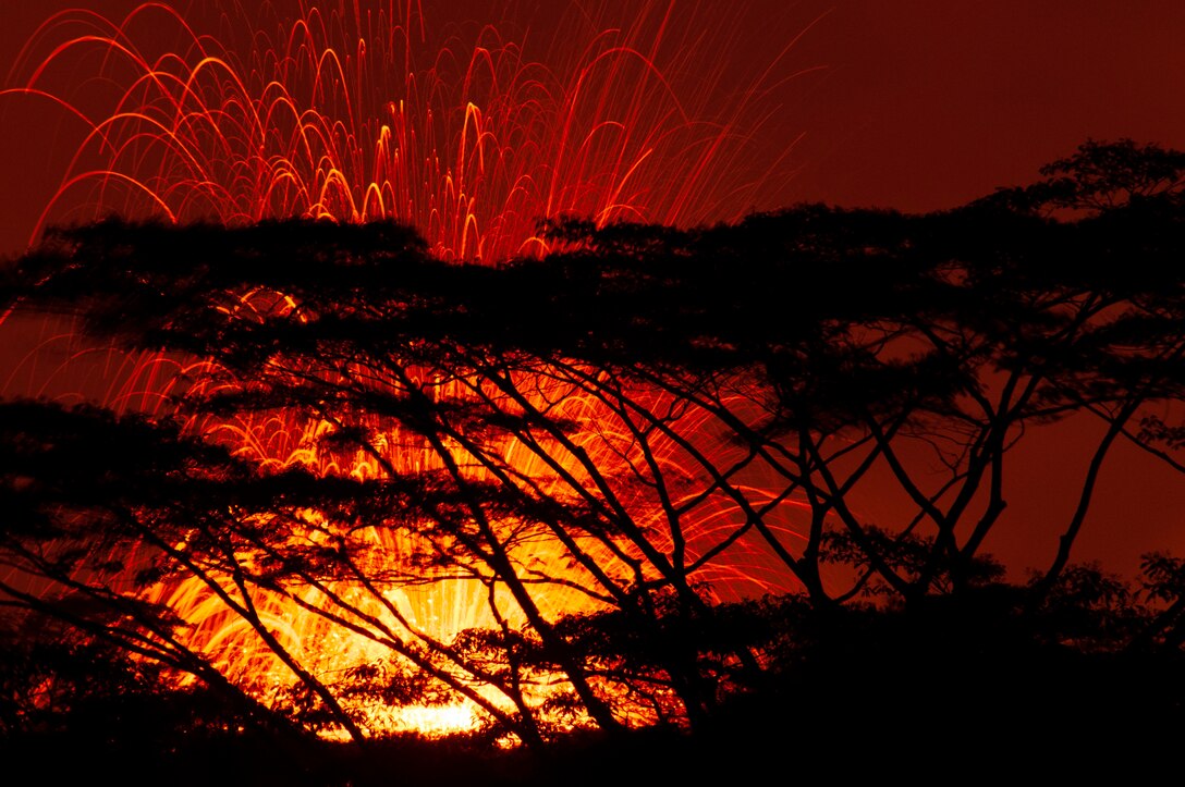 Flames and sparks shoot into the air, turning the sky red and orange behind silhouetted trees.