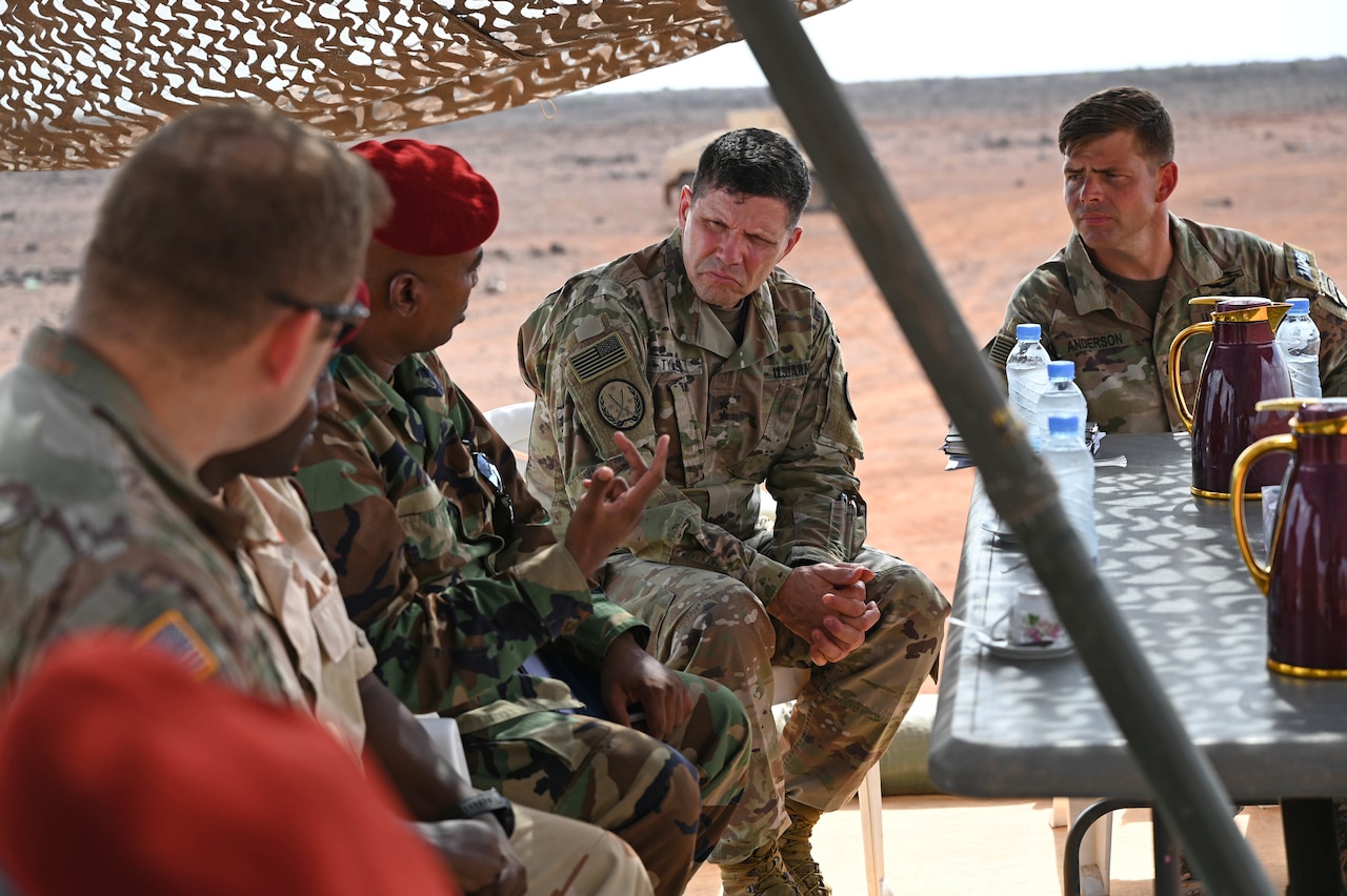 Shaded by an umbrella, five men in military uniforms sit at a table outside and talk; a desert landscape is in the background.