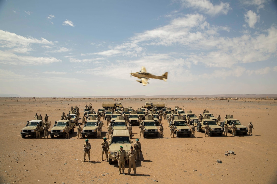 An aircraft flies over foreign military vehicles and service members in the desert.