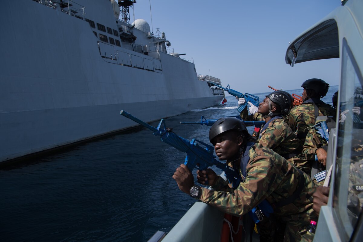 From the deck of a ship, service members point guns upward at a large ship.