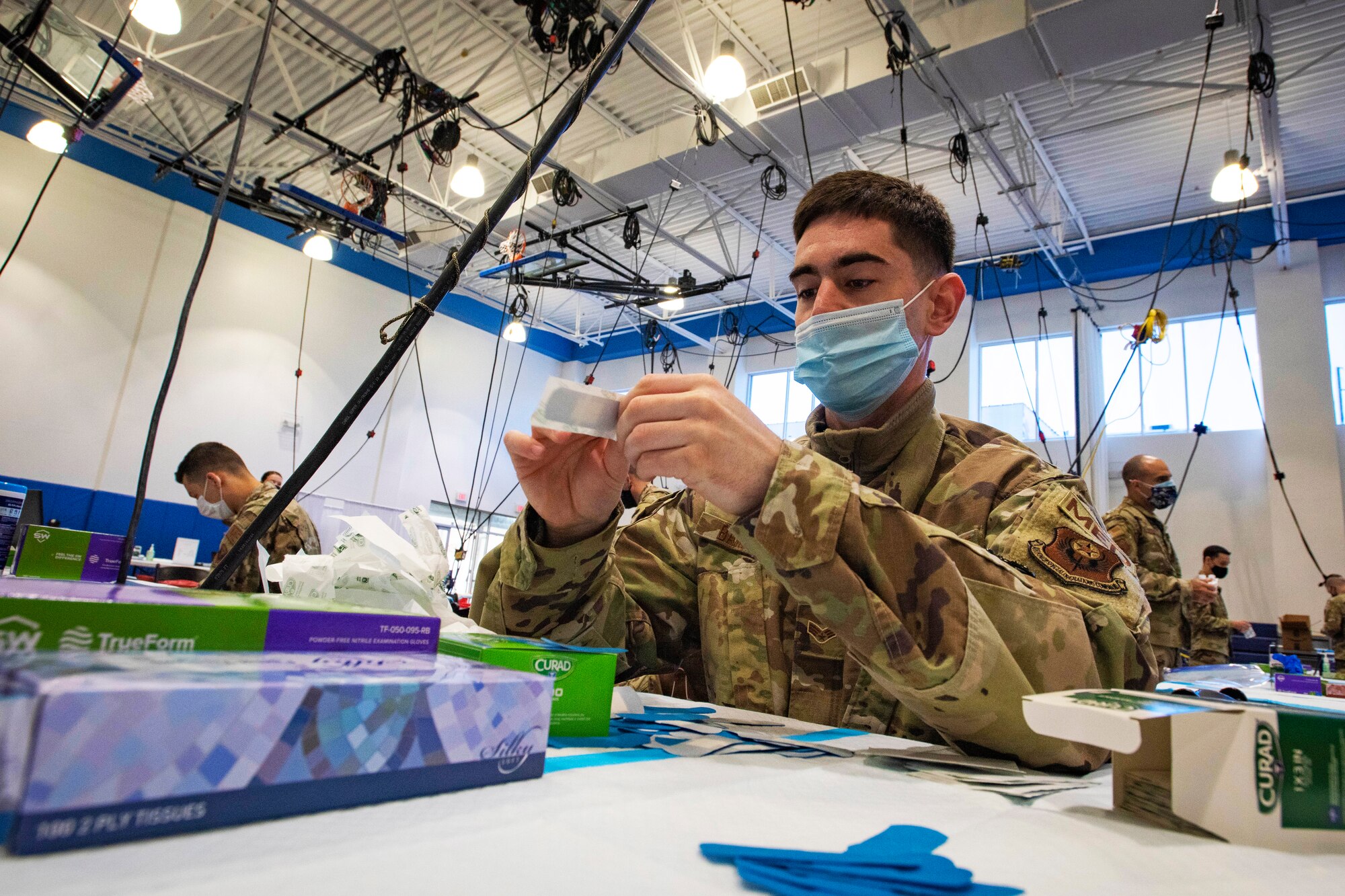 A soldier wearing a face mask sits at a table while opening a band-aid.