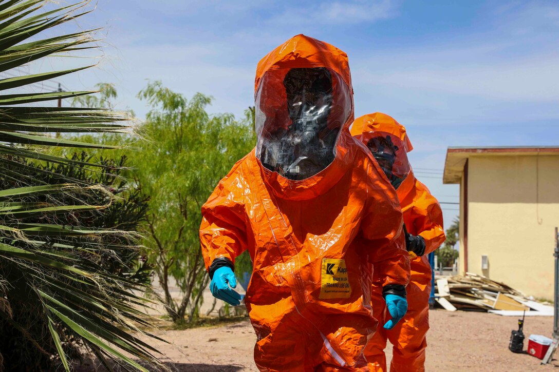 Two soldiers walk through desert terrain wearing orange hazmat suits.