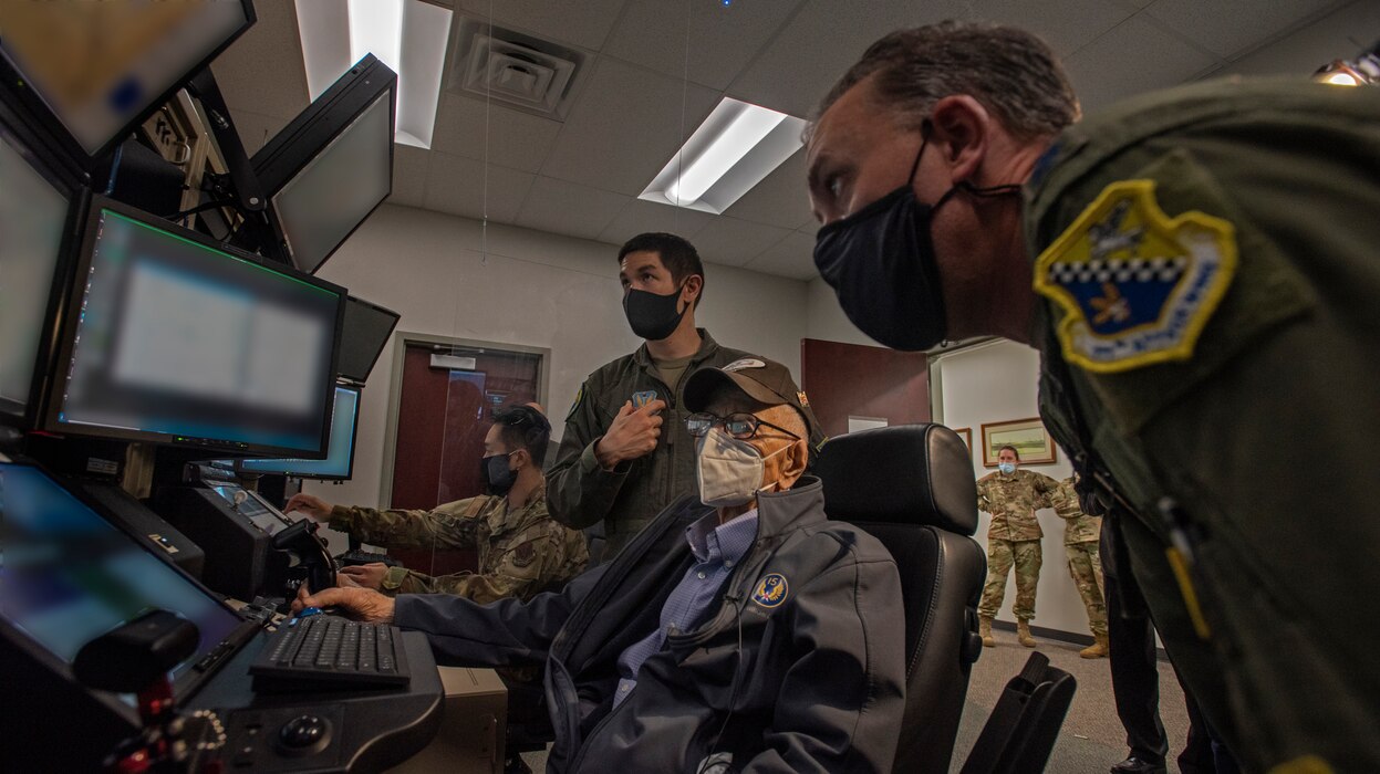 Two Air Force pilots in flight suits help an old man operate a flight simulator.