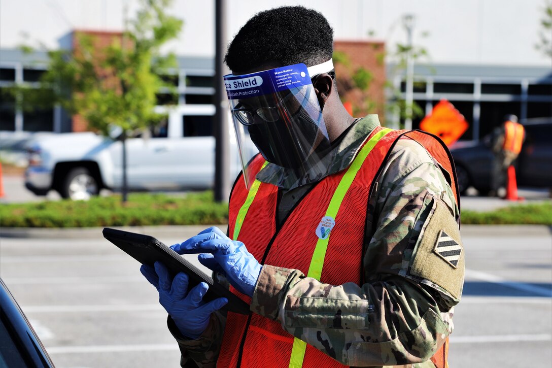 A soldier wearing personal protective equipment stands in a parking lot and works on an electronic tablet.