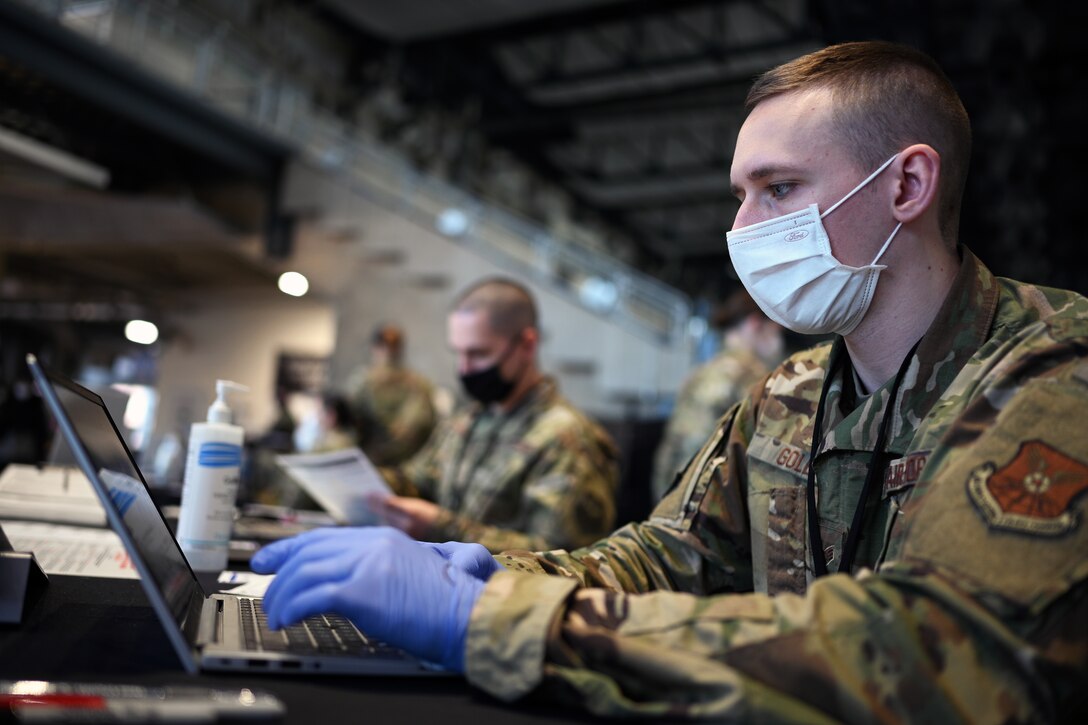 Several airmen sit at a table wearing face masks and gloves and work on laptops.