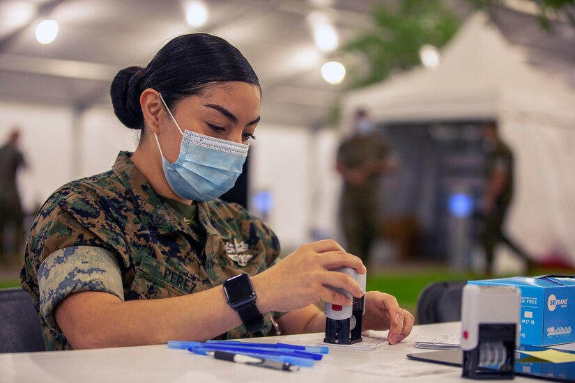 A sailor seated at a table and wearing a face mask stamps a small card.