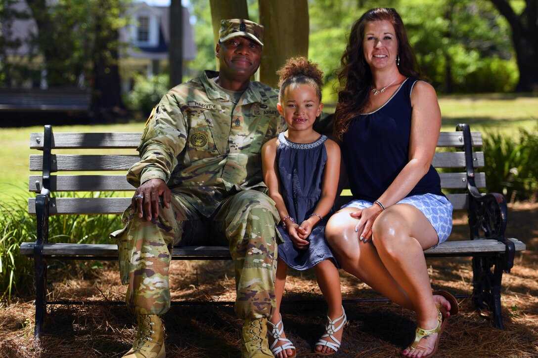 A soldier and his wife and daughter smile while sitting together on a park bench.
