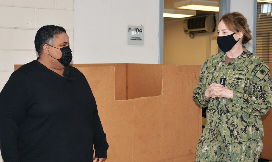A black woman in a black shirt and white woman in camo uniform - both wearing black face masks - stand facing each other in front of a cardboard box.