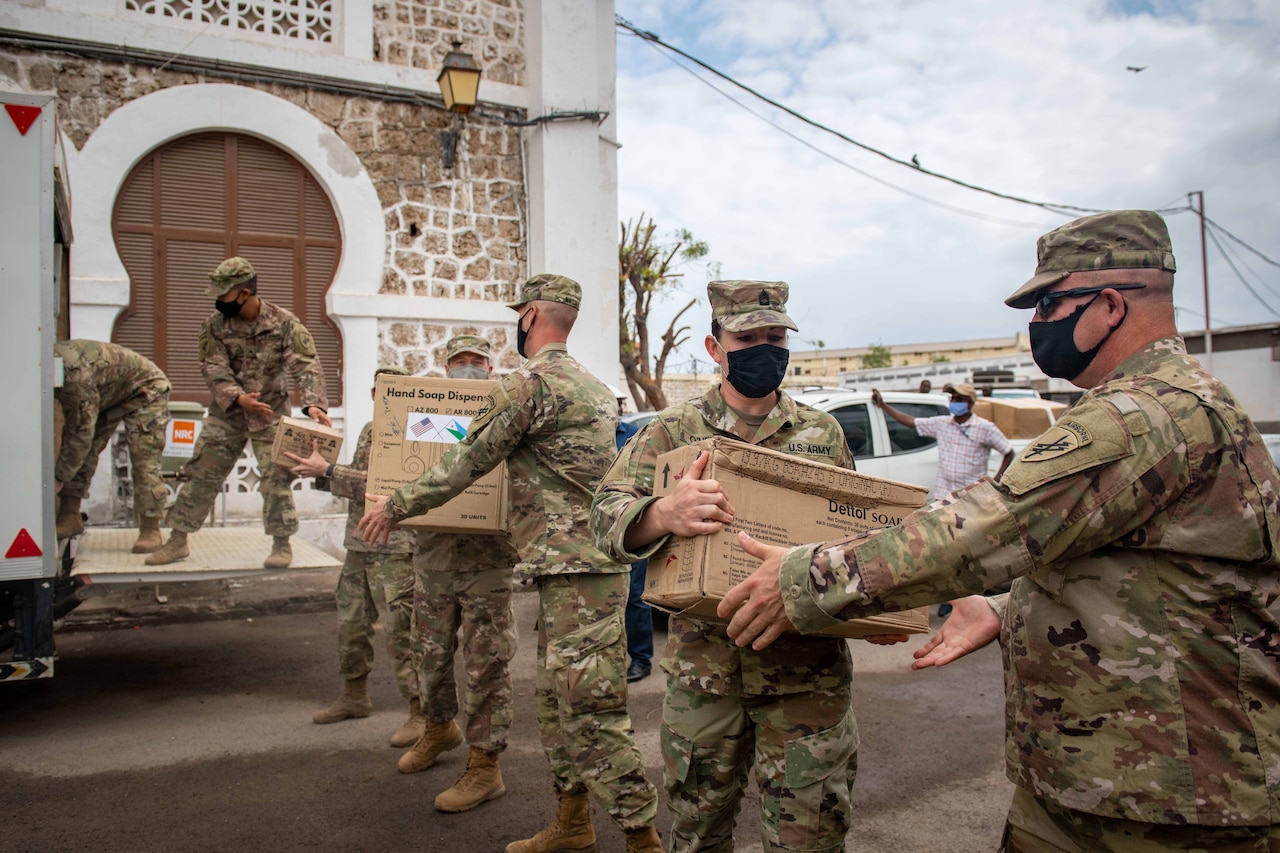 Soldiers in camouflage uniforms and face masks form a human chain to transfer large cardboard boxes.