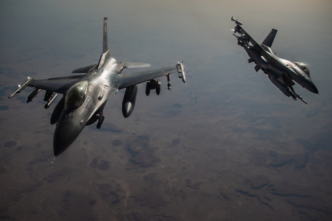 Jet fighters fly over a mixed terrain of farms and hills.