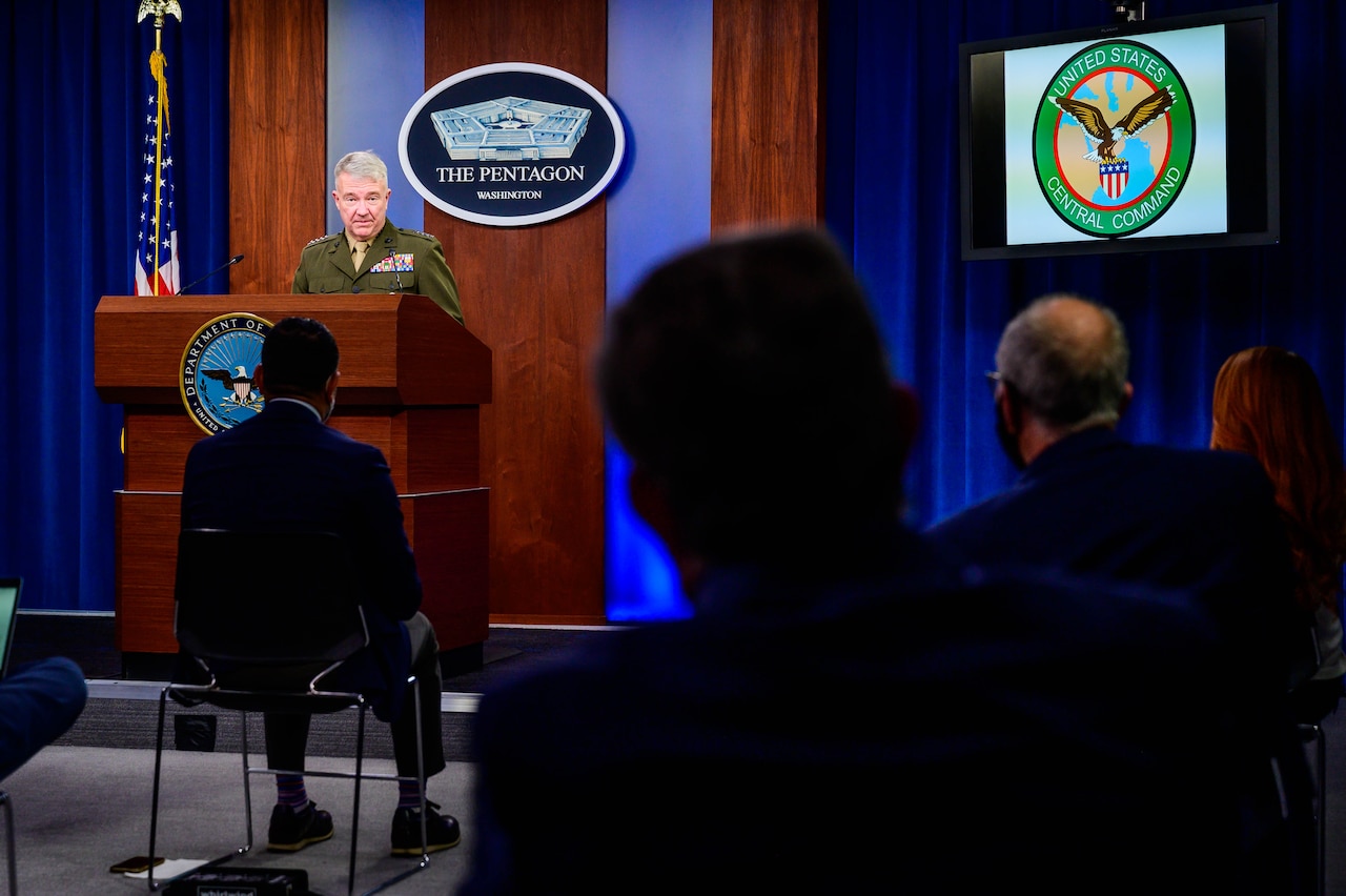 A man in a military dress uniform stands at a lectern. Behind him is a plaque indicating that he is at the Pentagon and seated reporters are silhouetted in the foreground.