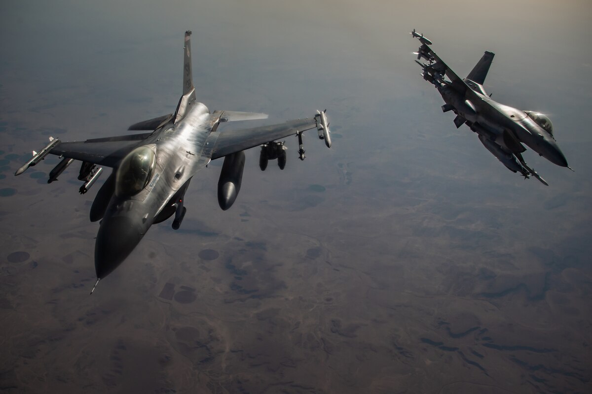 Jet fighters fly over a mixed terrain of farms and hills.
