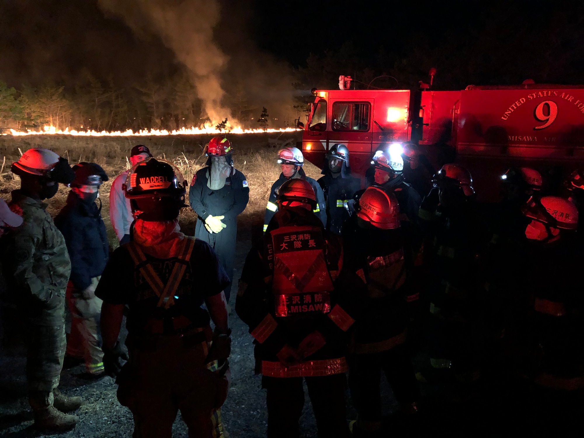 Fire crews stand near a fire truck and fire in a field