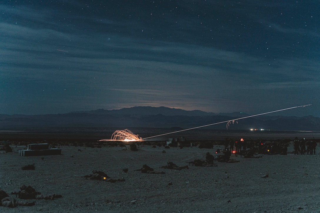 Marines lay on ground holding weapons in the desert while sparkly fly in the air nearby.