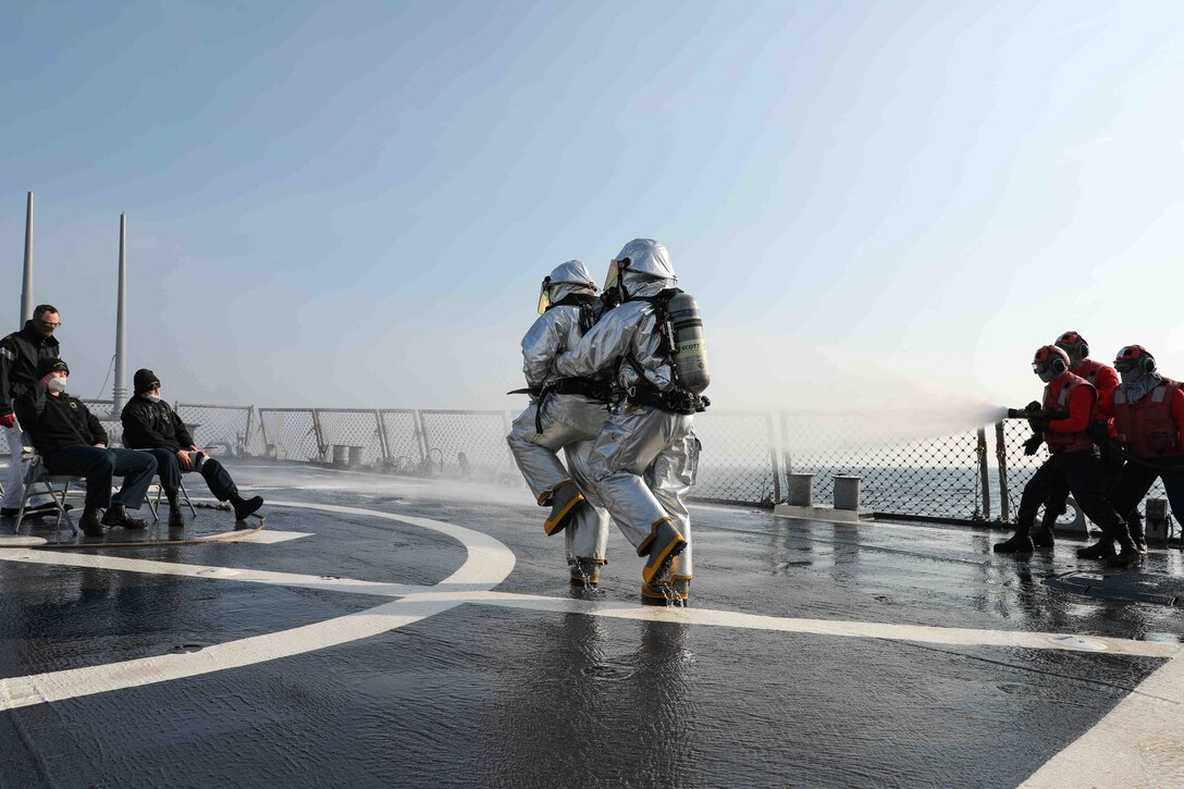 Sailors use a water hose to put out a simulated fire while other sailors wear fire suits on the flight deck of a ship at sea.