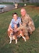 A man in uniform hugs his son. Their dog sits in front of them.