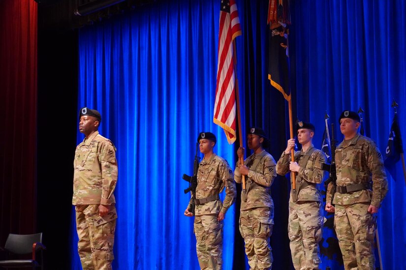 A soldier stands at attention in front of a color guard carrying the American flag.
