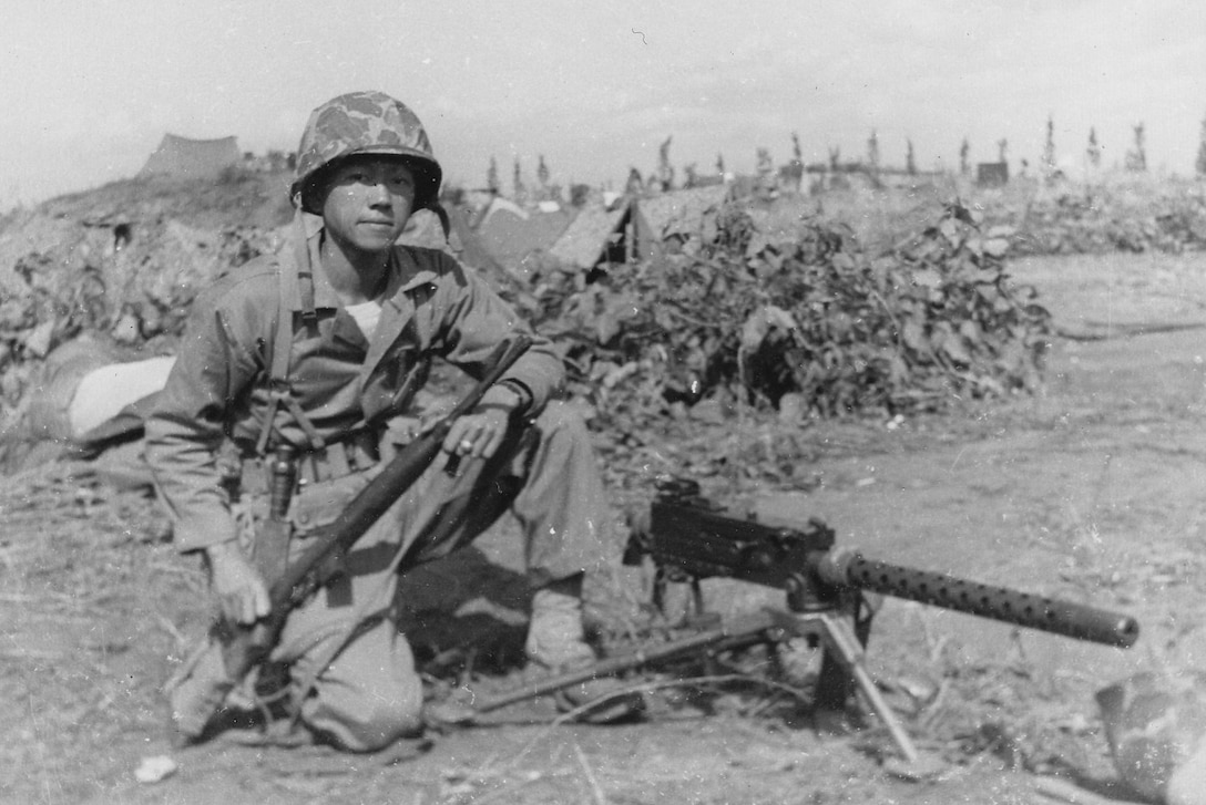 A Marine kneels outside by a machine gun set up on a stand.