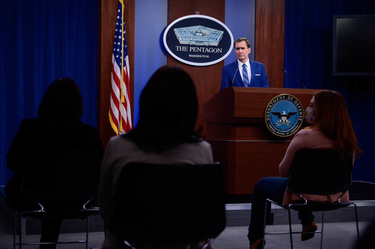 A man stands behind a lectern. Behind him is a sign that reads "The Pentagon -- Washington." In front of him are seated individuals.