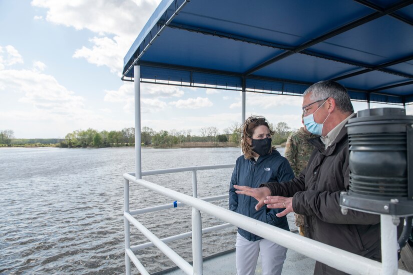 A man and woman speak to each other while standing near a safety rail and under an awning on a boat.