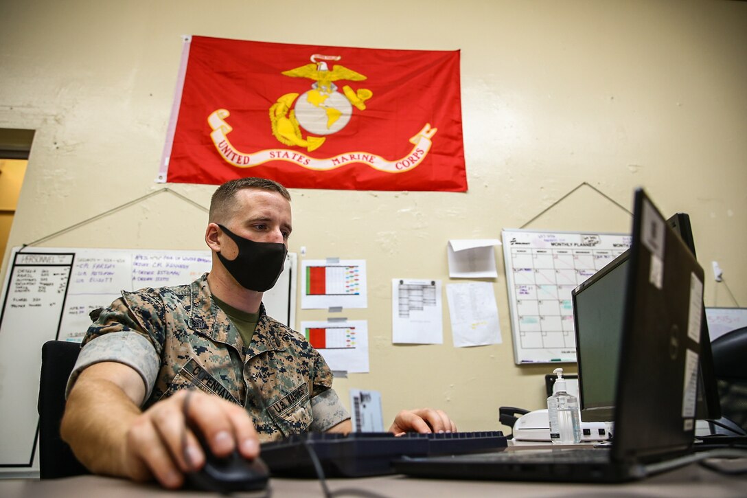 A Marine sits at his desk, holding his mouse, looking at a computer. The Marine Corps flag and other organizational posters are on the wall.