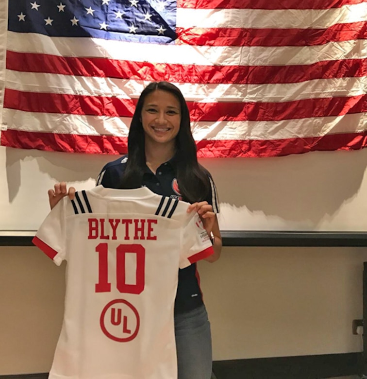 A woman standing in front of an American flag holds up her USA Rugby team jersey.