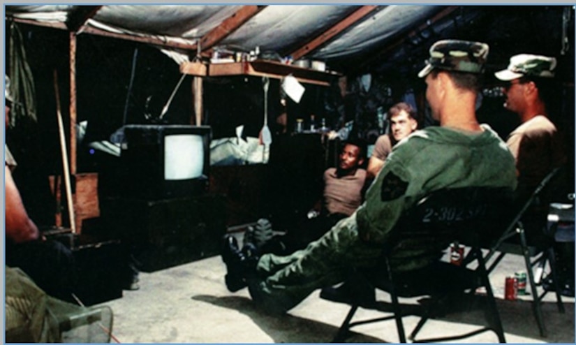 Five service members crowd around a small television in a tented room.