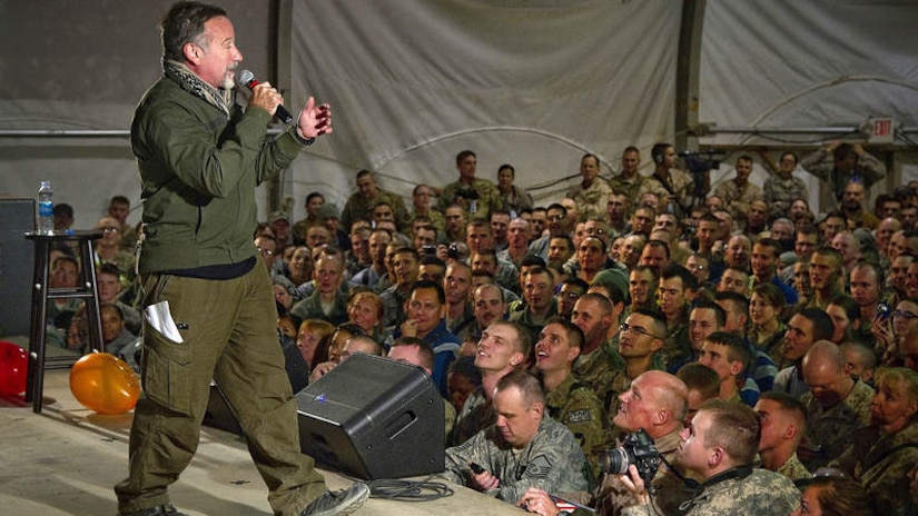 A man holds a microphone on stage while uniformed service members watch from below.