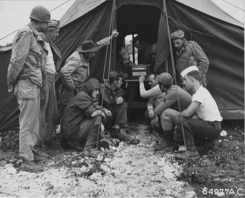 Several men crowd around a 1940s-era radio sitting just inside a tent.