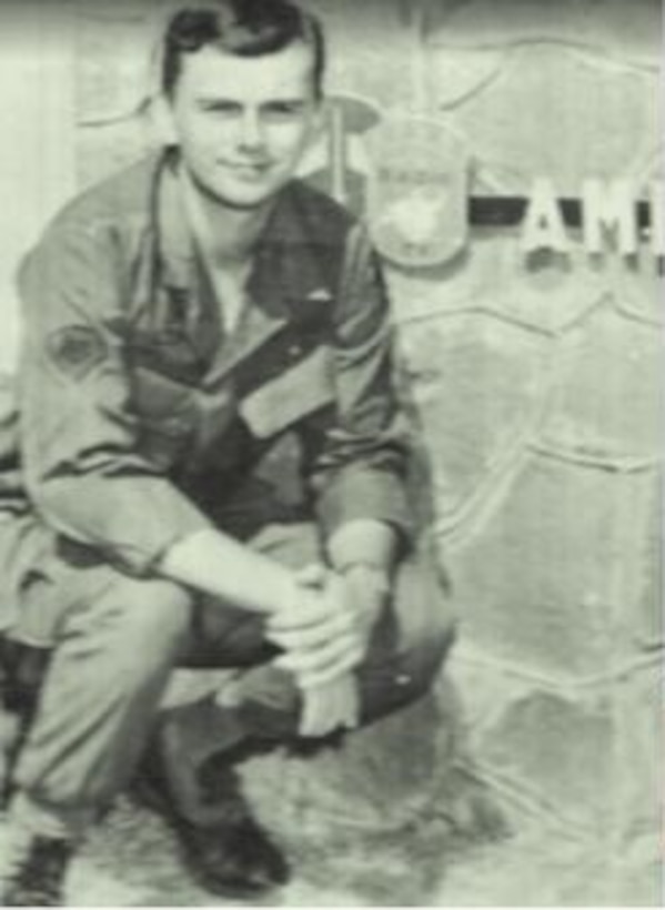 A man in uniform squats down to pose near a sign that's cut off.