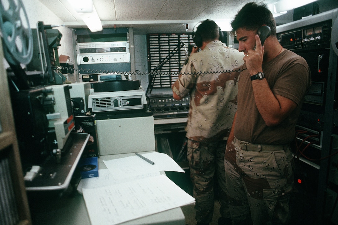 A man in fatigues is on the phone while another man dons headphones in a small studio.