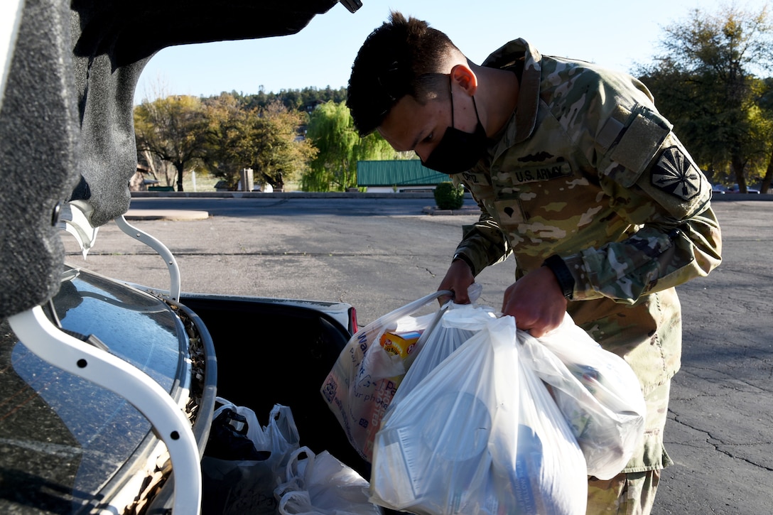 A male soldier wearing a face mask puts bags of groceries into the trunk of a car.