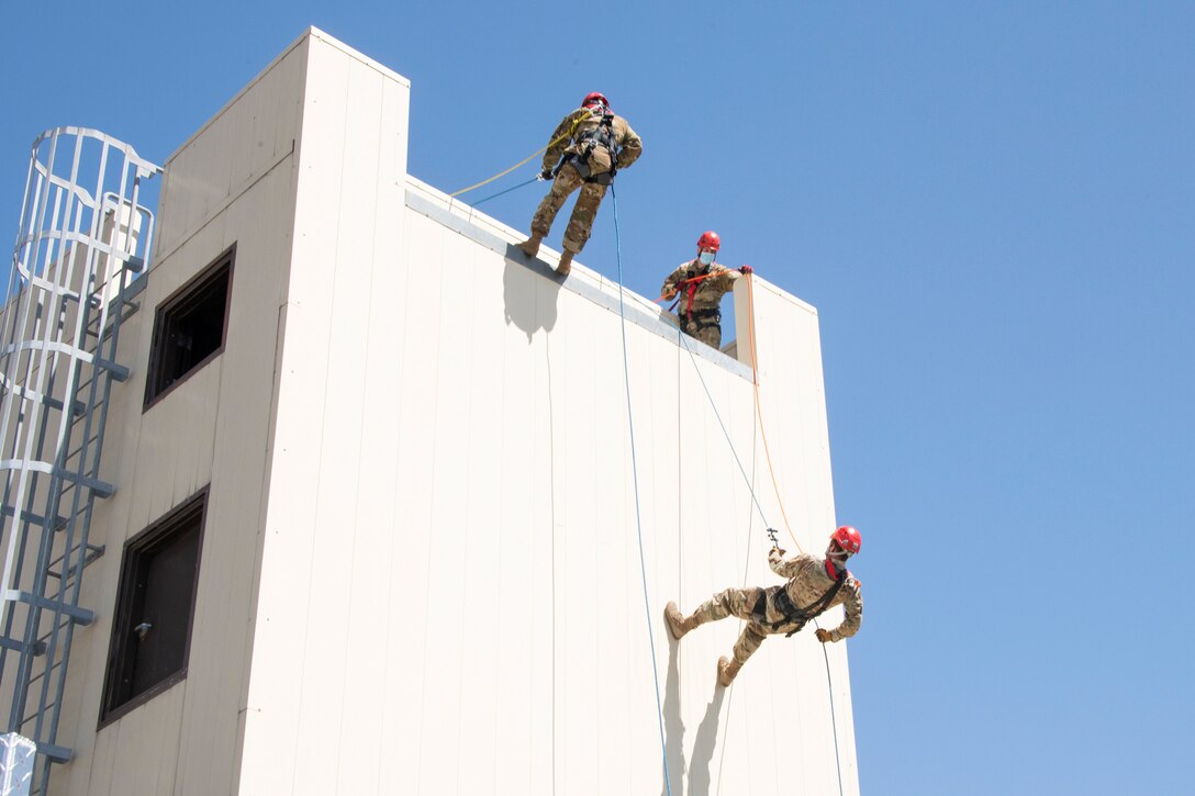 Two airmen rappel down a structure while a fellow airman watches.