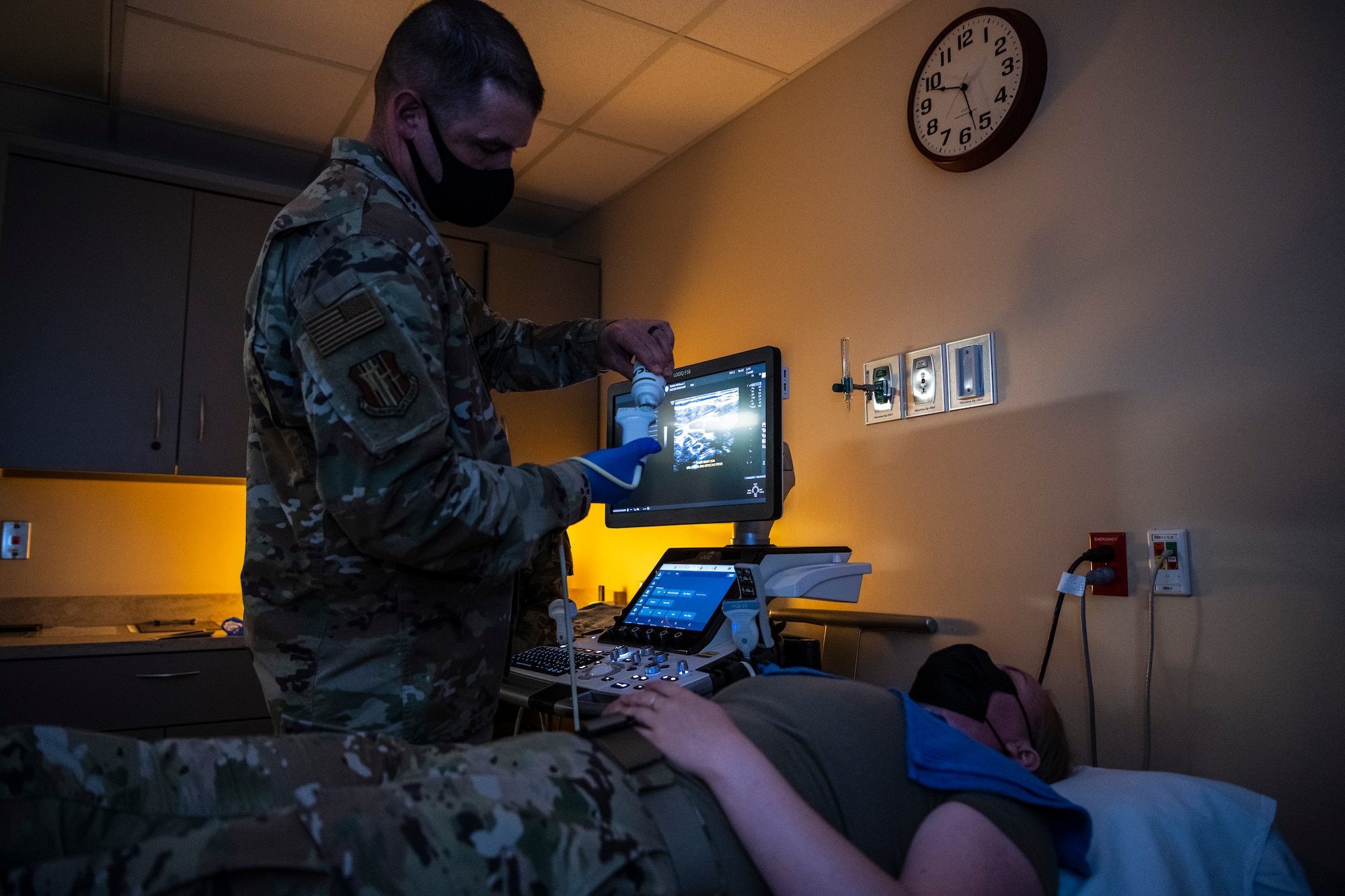 An Airman lays on a hospital bed as another Airman with a lot of rank is taught how to do an ultrasound to her neck.