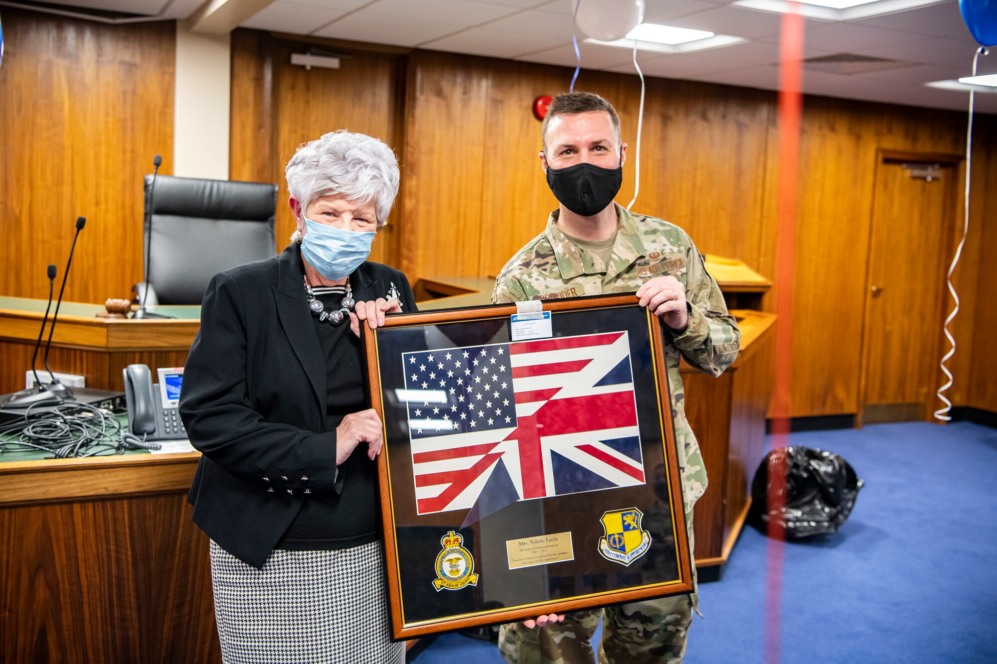 U.S. Air Force Maj. Richard Schrider, right, 501st Combat Support Wing staff judge advocate, presents a gift to Valerie Lucas,  501st Combat Support Wing British liaison officer, during a celebration at RAF Alconbury, England, April 23, 2021.  The 501 CSW/JA office celebrated Mrs. Lucas for her over 60 years of service as the British liaison officer at RAF Alconbury. (U.S. Air Force photo by Senior Airman Eugene Oliver)