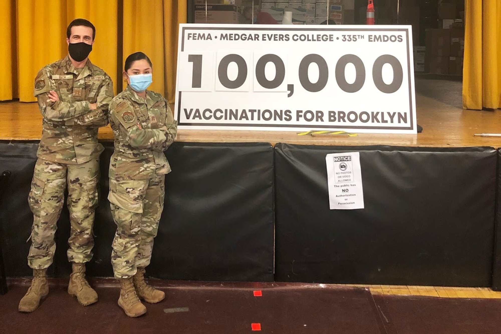 Two Air Force members stands in front of a sign reading 100,000 vaccinations for Brooklyn.