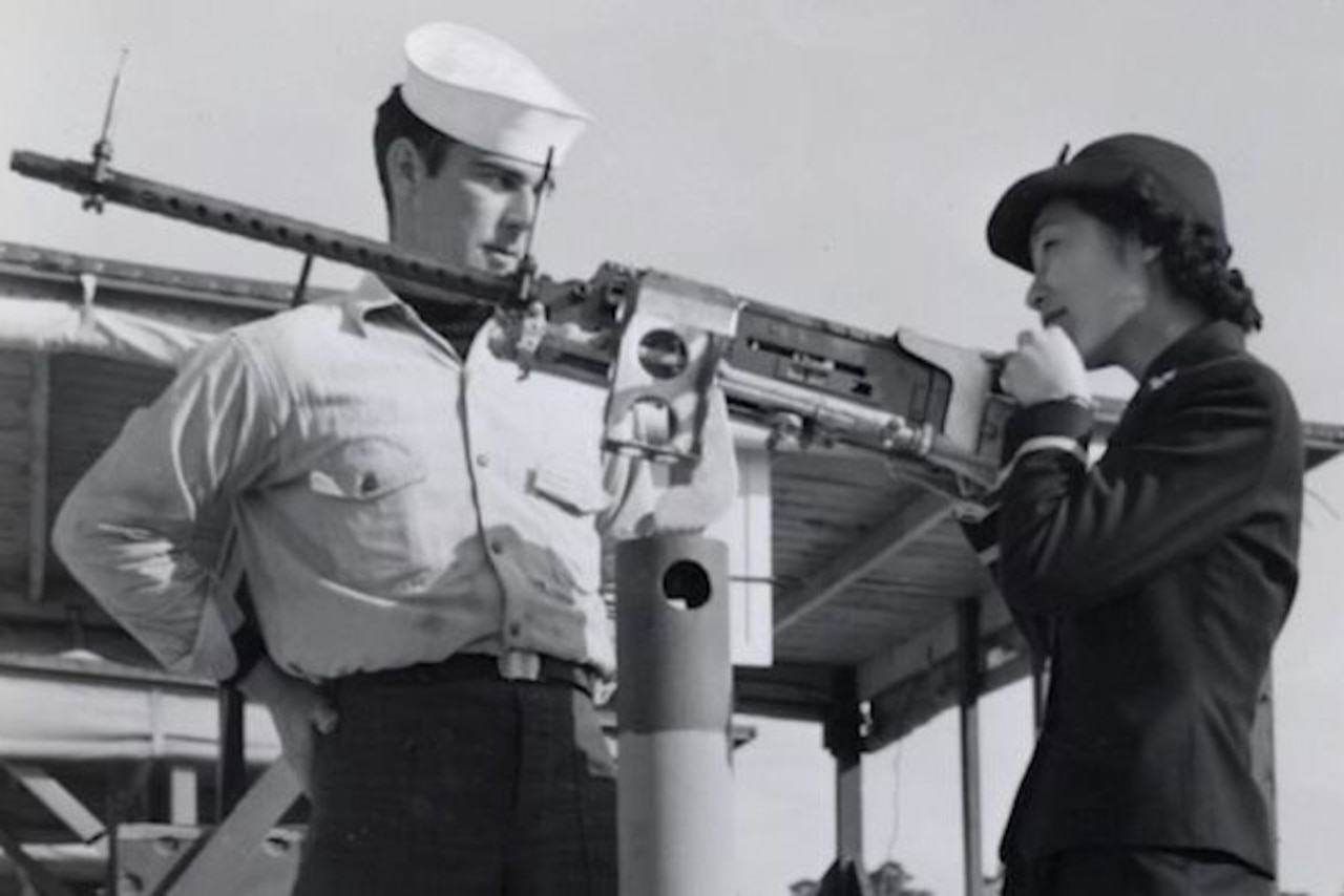 A man in uniform watches a woman in uniform as she looks through the scope of a long gun.