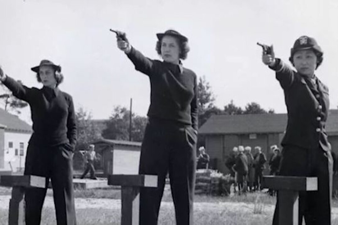 Women in a line aim pistols into the distance. Each has her left hand in her pocket. Others watch from a distance.