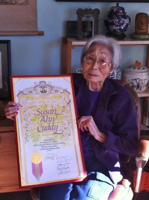 A woman holds up a plaque that says "Susan Ahn Cuddy."