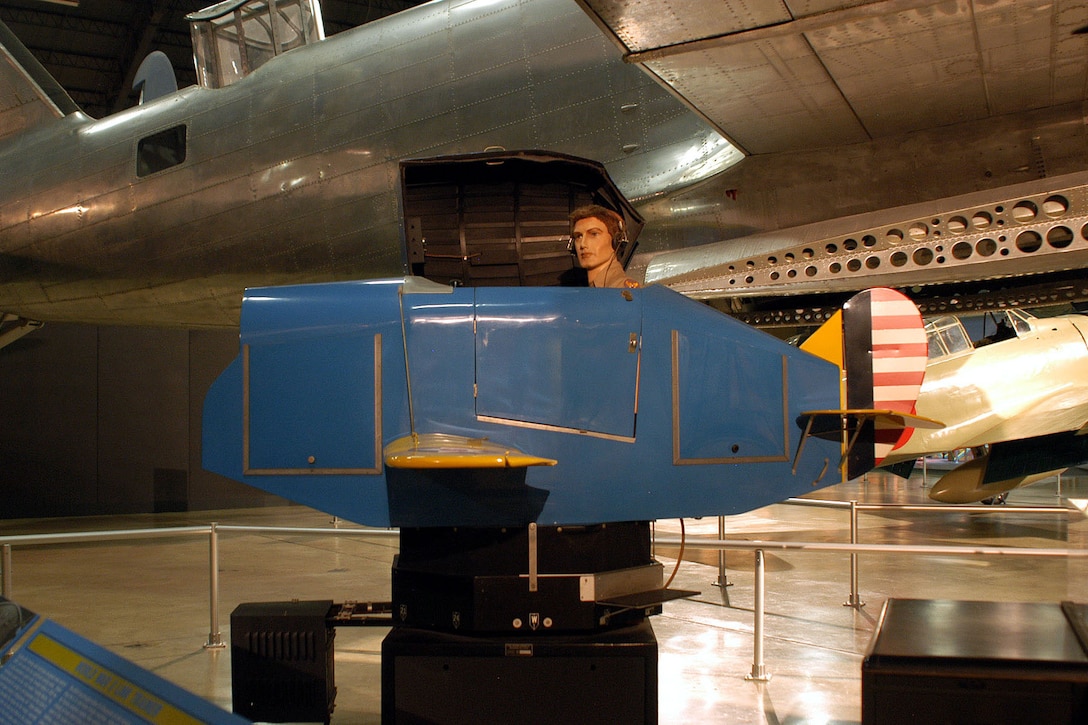 A mannequin's head pokes out from a small airplane simulator in a hangar with other aircraft.