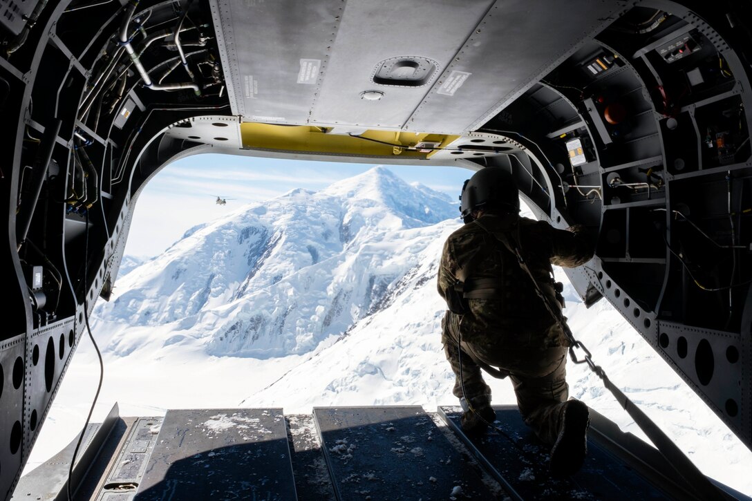 A soldier kneels on the ramp of an aircraft while looking at a snowy mountain.
