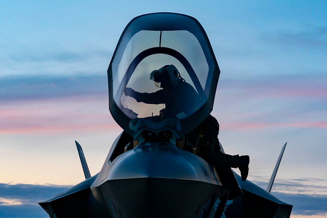 A Marine stands perched on an F-35B II Lightning II with an open cockpit.'
