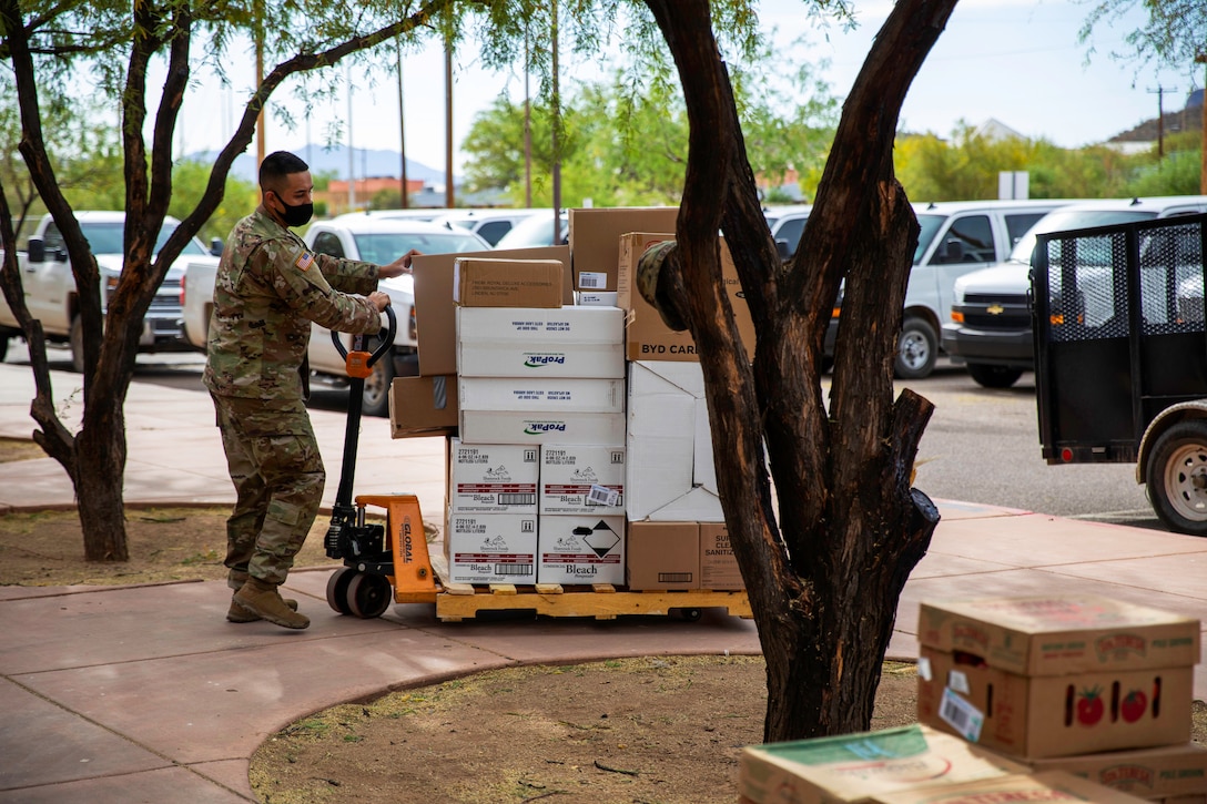 A soldier wears a facemask while moving a pallet stacked with boxes.